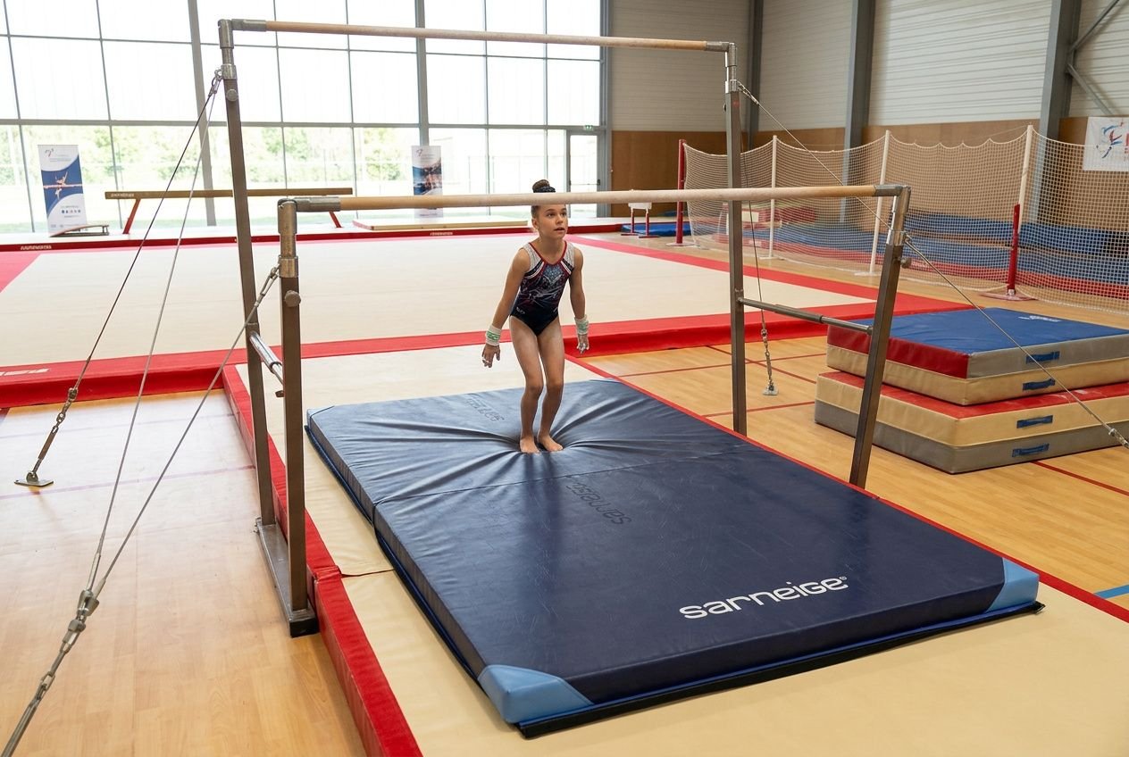 Petite gymnaste debout sur un grand matelas bleu, prêt pour la barre asymétrique gym dans une salle d’entraînement équipée.